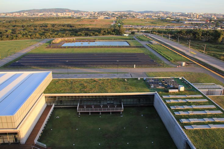 “Vista aérea de um complexo arquitetônico sustentável com telhados verdes, painéis solares, áreas ajardinadas e vias ao redor, integrado a uma paisagem urbana ao fundo.”