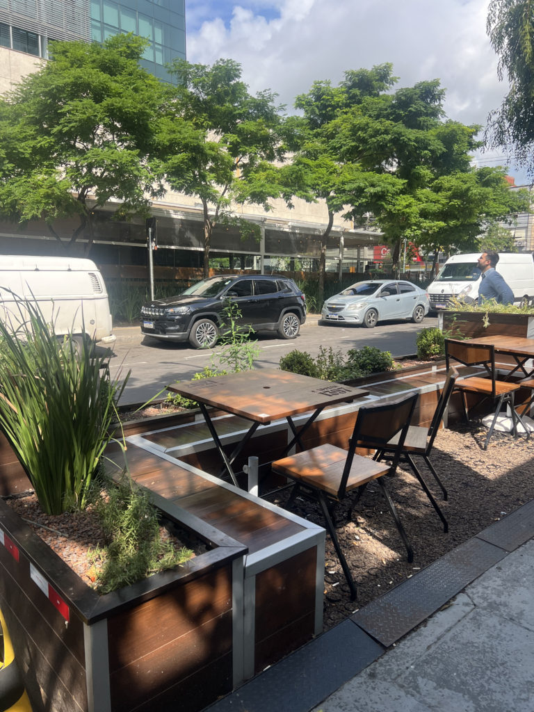 Área externa de restaurante com parklet urbano sustentável, equipada com mesas e cadeiras de madeira integradas a floreiras com vegetação. O espaço ocupa parte da via pública, promovendo convivência e contato com a natureza em meio ao ambiente urbano, com árvores ao redor e veículos estacionados na rua.