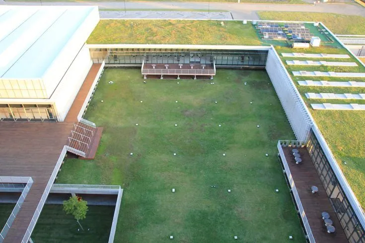 Vista aérea de um pátio interno de um edifício corporativo ou educacional coberto por um amplo gramado (telhado verde ou laje jardim). O espaço é cercado por passarelas de madeira e fachadas de vidro, mostrando uma grande área de infraestrutura verde urbana para controle térmico e lazer.