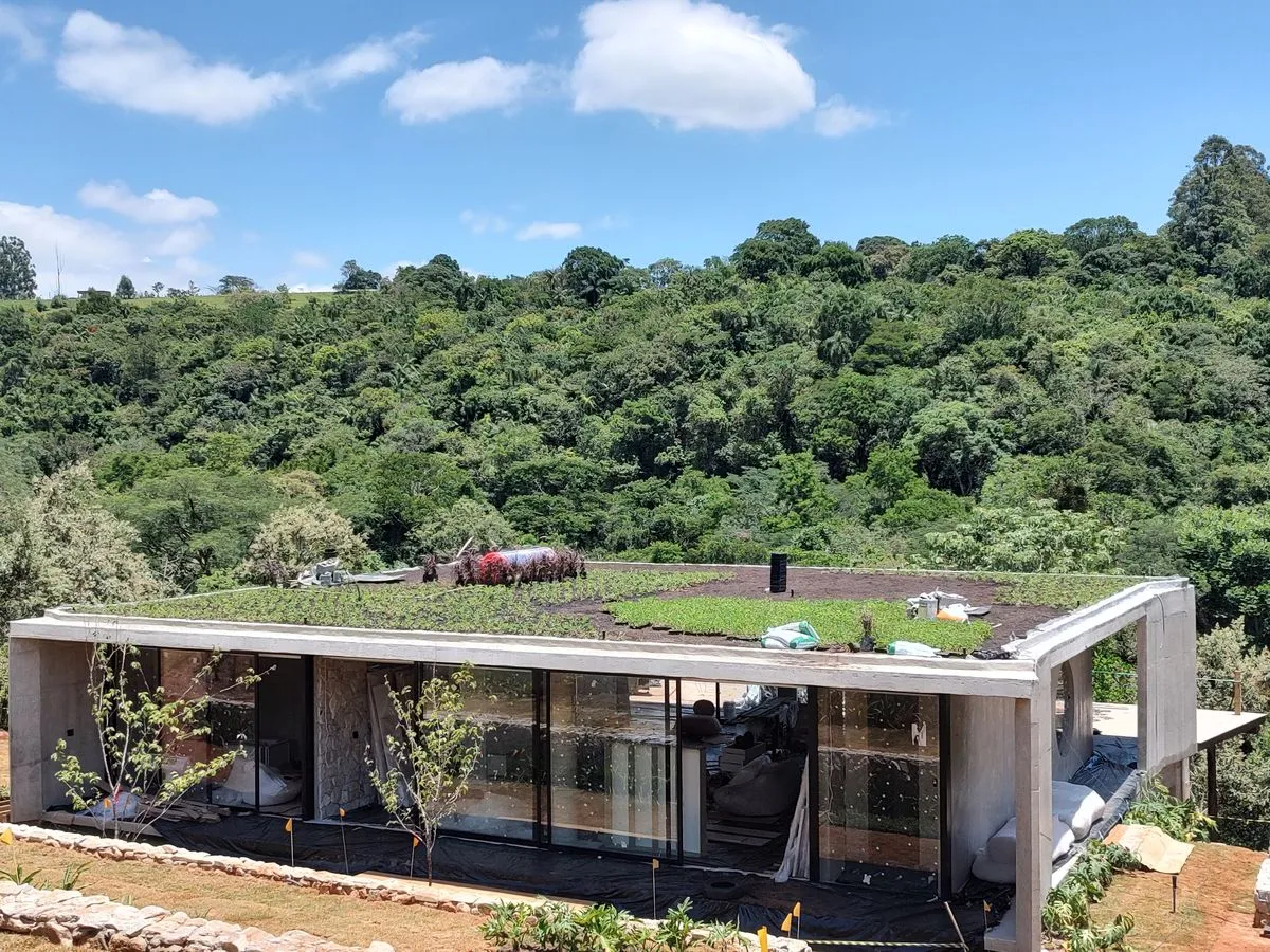 Vista externa de uma residência moderna de concreto em construção, destacando um amplo telhado verde em fase de instalação sobre a laje. A casa está inserida em uma encosta com vegetação nativa densa ao fundo, ilustrando a aplicação de soluções baseadas na natureza na arquitetura.