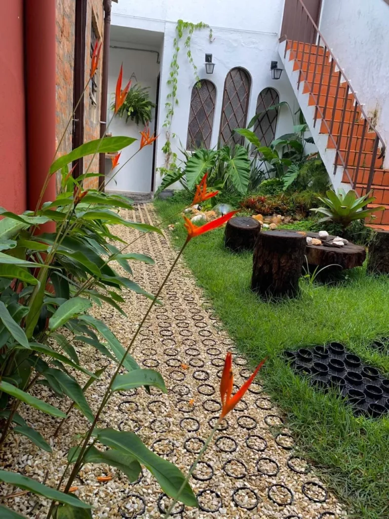 Caminho de jardim feito com ecopavimento preenchido por brita clara, cercado por gramado verde e plantas tropicais como Helicônias. O percurso leva ao pátio de uma edificação branca com detalhes em madeira e bancos de tronco de árvore, exemplificando o design biofílico e pavimentação permeável.