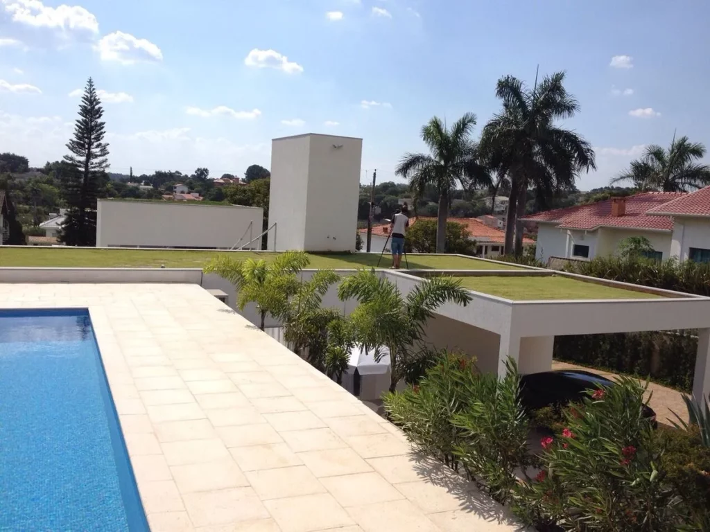 Vista aérea de uma residência de alto padrão com design moderno. O terraço sobre o nível inferior e o telhado da garagem são totalmente cobertos por um gramado verde e bem cuidado, funcionando como telhados verdes que se integram à paisagem e auxiliam no isolamento térmico e na gestão da água da chuva.