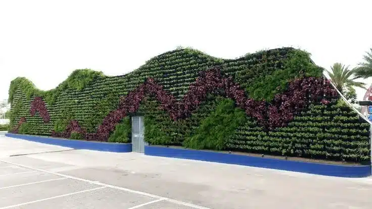 Uma longa e alta parede de jardim vertical com formato ondulado e orgânico ao lado de um estacionamento. O painel vegetal apresenta um design complexo e dinâmico, mesclando folhagens de cores verde vibrante e roxo profundo, destacando a capacidade de capturar poluentes e melhorar o microclima urbano.