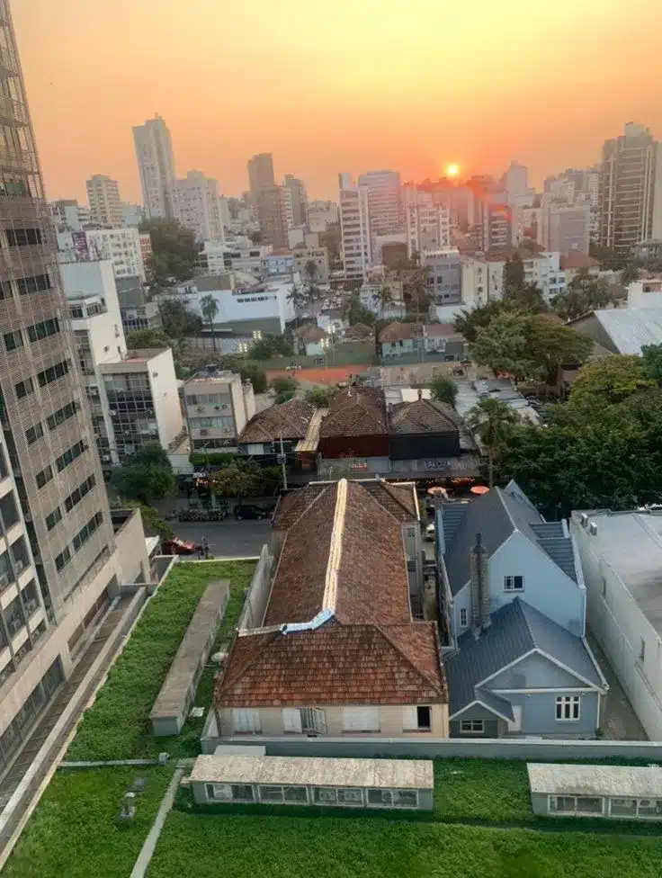 Vista aérea de um telhado verde em primeiro plano, que funciona como um regulador térmico e detém água da chuva. Atrás dele, o horizonte urbano de Florianópolis se estende ao pôr do sol, revelando a proximidade entre as edificações antigas e novas. O telhado verde exemplifica a integração da "quinta fachada" com a paisagem, conforme descrito no projeto, e contrasta com a arquitetura tradicional e os arranha-céus, evidenciando o esforço de reequilíbrio urbano e a conexão com a natureza defendida pelo design biofílico.
