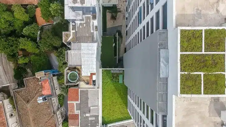 Vista aérea inclinada de um ambiente urbano denso em Florianópolis. À direita, um edifício moderno exibe dois grandes telhados verdes divididos em seções, como pilares da estratégia de sustentabilidade do projeto na Avenida Beira-Mar Norte. No centro, uma área gramada estreita conecta as edificações. À esquerda, as coberturas antigas de telhas de cerâmica da cidade histórica contrastam com a infraestrutura verde contemporânea, ilustrando a mudança de paradigma para uma cidade mais biofílica e resiliente, conforme as diretrizes do Gehl Architects.