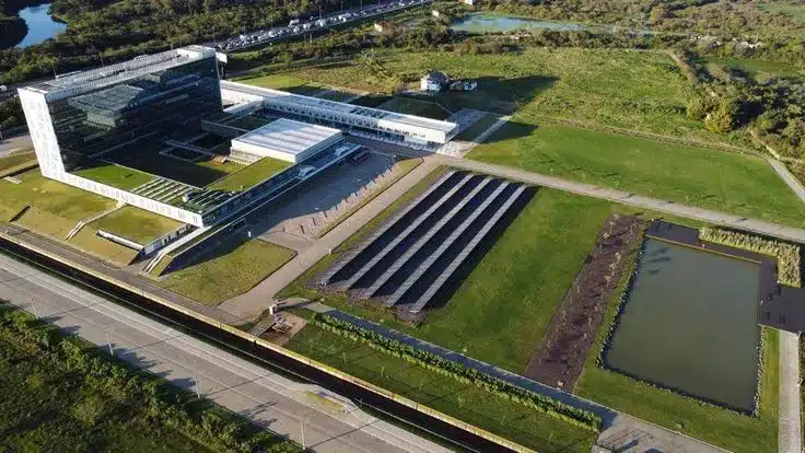 ista aérea ampla de um complexo arquitetônico moderno e sustentável à beira d'água, representando o Parque Urbano Marina na Beira-Mar Norte. O grande edifício em forma de L e as estruturas mais baixas em seu pátio interno apresentam telhados vegetados e paredes vegetadas, exemplos de design biofílico. No primeiro plano, uma ampla bacia de detenção d'água e áreas gramadas com drenagem sustentável demonstram o compromisso do projeto com a gestão de águas pluviais, infiltração e resiliência ambiental para a Baía Norte.