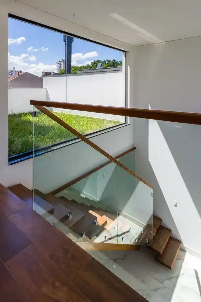 Vista aérea interna inclinada de uma escadaria moderna de dois lances com degraus e corrimão de madeira e guarda-corpos de vidro. No primeiro plano, o patamar superior exibe o início da descida. No nível intermediário, uma grande janela do chão ao teto oferece uma vista para um telhado verde com vegetação rasteira, que funciona como um regulador térmico e detém água da chuva. O telhado verde exemplifica a integração da "quinta fachada" com a paisagem, conforme descrito no projeto, e contrasta com a arquitetura tradicional e os arranha-céus, evidenciando o esforço de reequilíbrio urbano e a conexão com a natureza defendida pelo design biofílico. No fundo, a cidade de Florianópolis se estende sob um céu azul com nuvens brancas. No nível inferior, a base da escadaria termina em um piso de mármore branco com veios cinza.