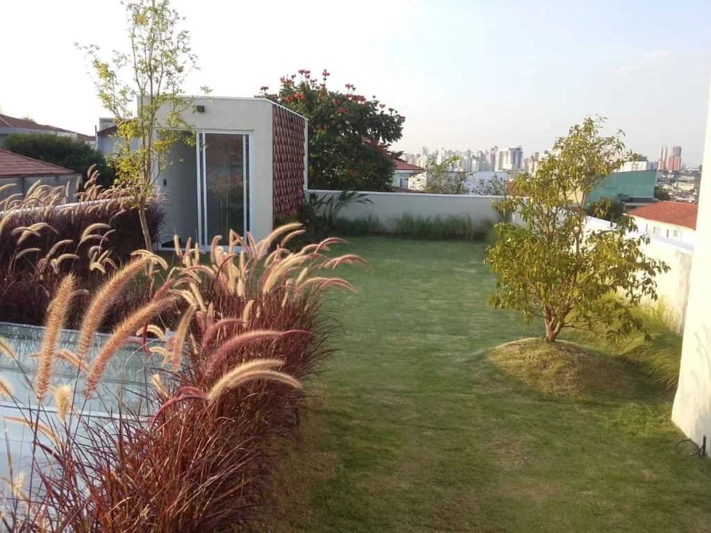 Terraço ajardinado com gramado verde, árvores de pequeno porte e capim-do-pêlo (Pennisetum setaceum) em destaque. O espaço de convivência externo demonstra como a infraestrutura verde serve como vantagem competitiva e hedge financeiro.