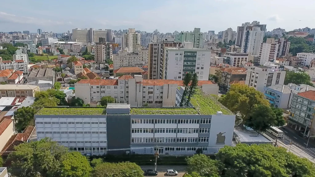 Vista aérea panorâmica do Colégio Israelita inserido no cenário urbano. Em primeiro plano, destaca-se a cobertura do edifício escolar em formato de "L", totalmente revestida por um extenso telhado verde gramado com cerca de 1.000 m². Duas árvores altas de copa estreita sobressaem do teto vivo. O prédio possui fachada cinza e branca repleta de janelas alinhadas. Ao redor, há árvores frondosas no nível da rua e, ao fundo, estende-se uma densa paisagem urbana com diversos edifícios residenciais e comerciais sob um céu parcialmente nublado.