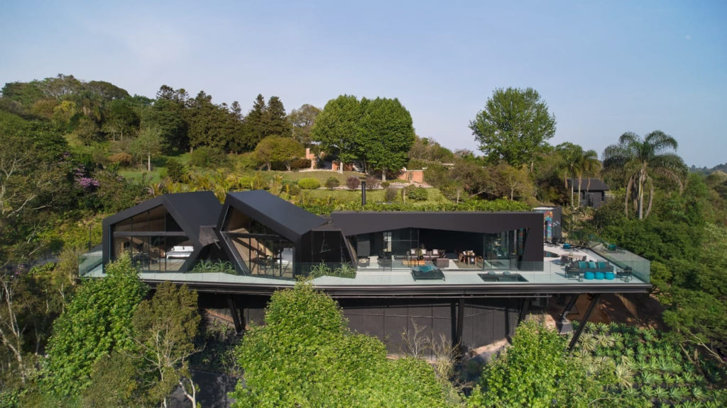 Vista aérea de uma residência contemporânea de alto padrão integrada à natureza. A casa possui uma estrutura linear suspensa com fachada preta e grandes panos de vidro que revelam o interior iluminado e decorado. O teto apresenta linhas geométricas assimétricas e angulares. Na frente, há uma extensa varanda com guarda-corpo de vidro, abrigando espreguiçadeiras e sofás em tons de azul e cinza. A edificação está cercada por uma densa vegetação nativa, com árvores altas ao fundo e plantas ornamentais cobrindo o terreno em declive abaixo da estrutura.