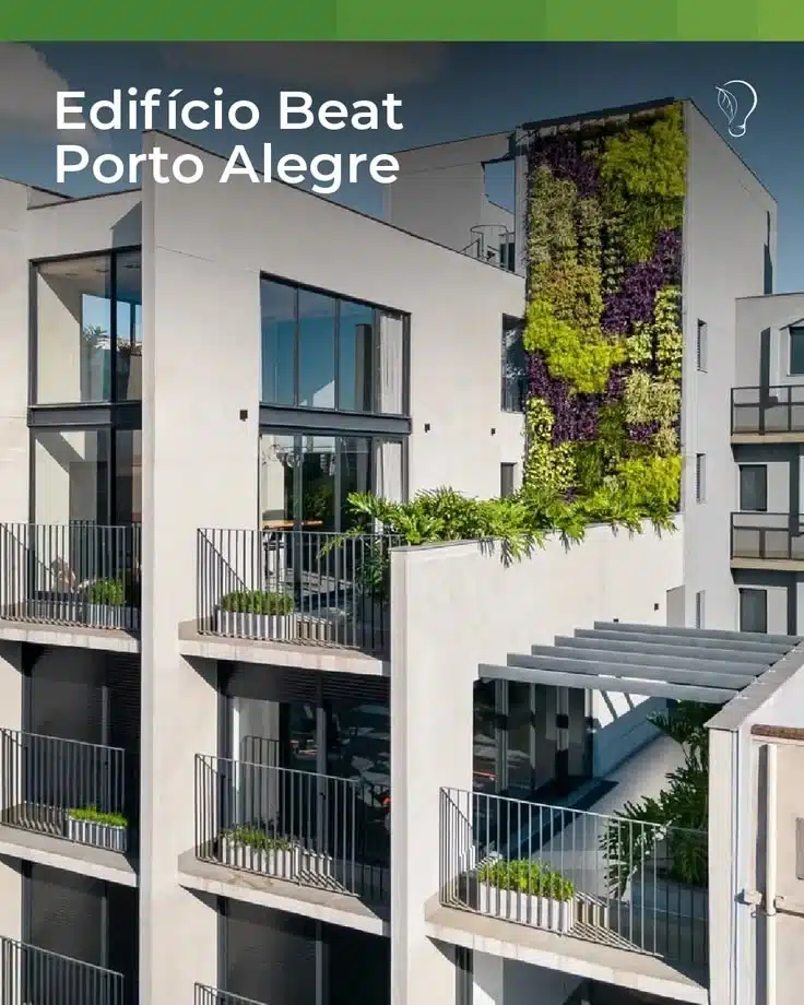 Fotografia em ângulo contra-plongé mostrando a fachada externa do "Edifício Beat", localizado em Porto Alegre, que serve como exemplo prático de bioarquitetura e urbanismo sustentável. O prédio possui um design contemporâneo e minimalista, com estrutura de concreto em tons de cinza-claro, grandes janelas de vidro com esquadrias escuras e sacadas lineares com guarda-corpos metálicos pretos vazados. Em cada sacada, encontram-se floreiras retangulares com pequenas vegetações.
O grande destaque visual da imagem é um imenso jardim vertical instalado em uma das paredes externas mais altas do edifício. A parede verde exibe um mosaico denso e orgânico de folhagens com diferentes texturas e cores, incluindo tons de verde-limão, verde-escuro e roxo-fechado. Abaixo dele, em um terraço intermediário protegido por um pergolado cinza, há canteiros adicionais com plantas tropicais de folhas largas. No canto superior esquerdo, o texto em letras brancas identifica: "Edifício Beat Porto Alegre". A composição ilustra perfeitamente como os jardins verticais e as fachadas vegetadas reinserem a natureza em ambientes urbanos densos para melhorar a regulação térmica e o bem-estar psicológico das pessoas.