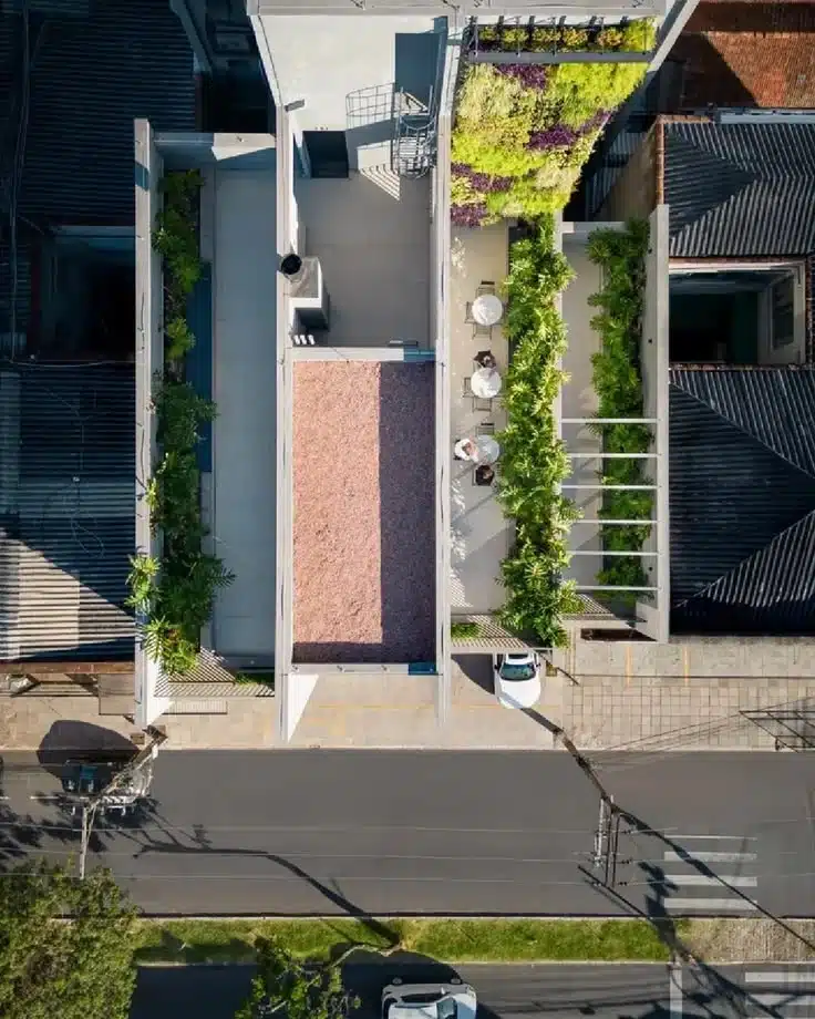 Vista aérea vertical (vista de drone) mostrando a cobertura e os terraços integrados do Edifício Beat em Porto Alegre. A imagem revela a inteligente ocupação do espaço horizontal por meio da infraestrutura verde e da bioarquitetura. No centro, uma das lajes superiores funciona como um telhado verde preenchido por substrato mineral de tom terracota.
À direita, um terraço de convivência exibe mesas brancas redondas com cadeiras dispostas ao longo de um corredor ladeado por densas fileiras de plantas tropicais verdes e um pergolado. Na extremidade superior desse mesmo lado, observa-se o topo do imenso jardim vertical multicolorido (com folhagens verdes, amarelas e roxas). À esquerda, outra seção de terraço linear também abriga canteiros vegetados. Na parte inferior, a fachada do prédio se alinha com a calçada e a rua asfaltada, onde carros estão estacionados. A imagem exemplifica visualmente o princípio do urbanismo biofílico discutido no artigo: a transformação de superfícies antes improdutivas em áreas ativas que retêm água da chuva, reduzem ilhas de calor e restabelecem a conexão humana com a natureza.