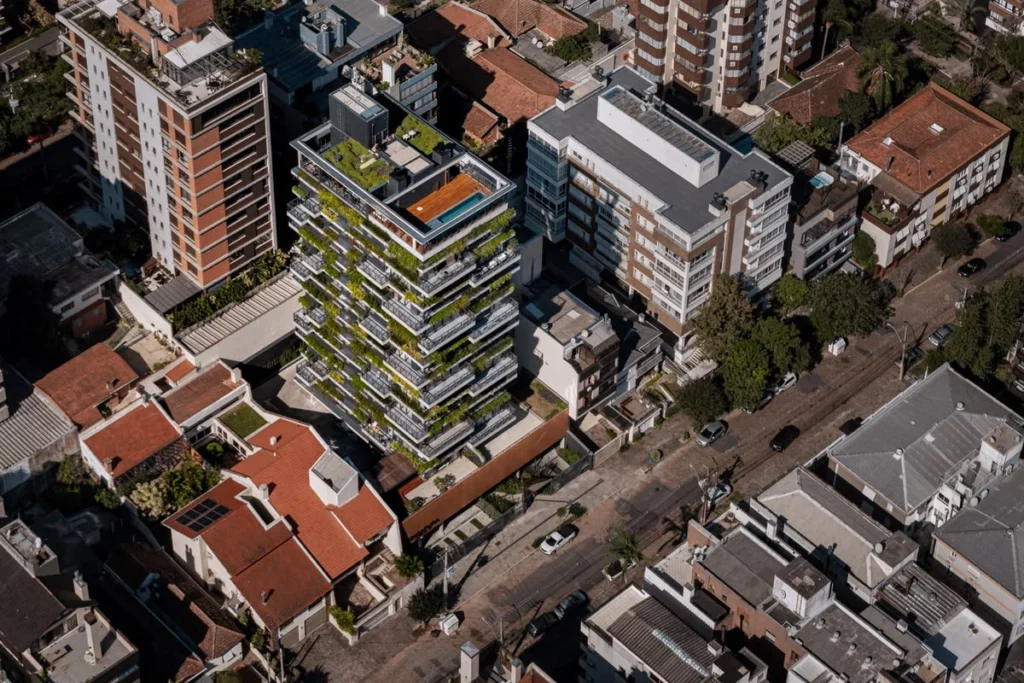 Fotografia aérea oblíqua mostrando um quarteirão residencial urbano densamente ocupado. No centro do plano, destaca-se um edifício residencial de múltiplos andares projetado sob as diretrizes da bioarquitetura e do urbanismo biofílico. O prédio apresenta varandas lineares em todos os andares, completamente cobertas por jardins suspensos e densas floreiras de vegetação verde-viva que envolvem a fachada.
Na cobertura (rooftop), observa-se um sistema completo de telhado verde com gramado natural, ladeando uma área de lazer com deque de madeira e uma piscina. O edifício sustentável contrasta diretamente com os prédios e casas convencionais ao redor, cujas coberturas expõem apenas telhas tradicionais de cerâmica e lajes cinzas de concreto. A imagem ilustra de forma clara o conceito do artigo sobre como a infraestrutura verde integrada transforma superfícies verticais e horizontais para combater o efeito de ilhas de calor e aumentar a resiliência climática das cidades.