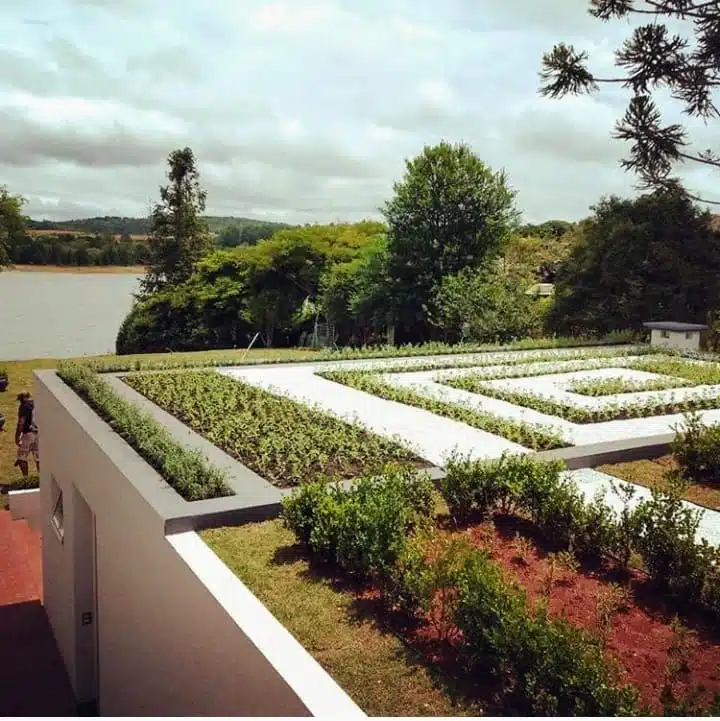 Fotografia em plano médio e ângulo oblíquo alto mostrando um amplo telhado verde projetado sobre uma edificação residencial ou comercial de linhas modernas e paredes brancas. A cobertura verde foi planejada como um jardim geométrico contemporâneo, apresentando canteiros lineares elevados e dispostos em formato de labirinto quadrado ou espiral retangular, onde crescem pequenas mudas de plantas e vegetações rasteiras organizadas. Os canteiros são intercalados por caminhos de pedriscos brancos.
No primeiro plano à direita, há uma seção de jardim convencional sobre terra avermelhada com arbustos verdes mais altos. Ao fundo, o cenário se abre para uma paisagem natural exuberante, margeada por uma grande lagoa ou rio à esquerda e árvores nativas densas ao centro, sob um céu parcialmente nublado. A imagem traduz perfeitamente o conceito do artigo sobre conceber empreendimentos desde o início integrados à topografia e à natureza, utilizando sistemas de telhados verdes tecnológicos (como as soluções laminares da Ecotelhado) que funcionam como bacias de detenção suspensas para reter a água da chuva, reduzir temperaturas e promover a eficiência energética e o conforto térmico.
