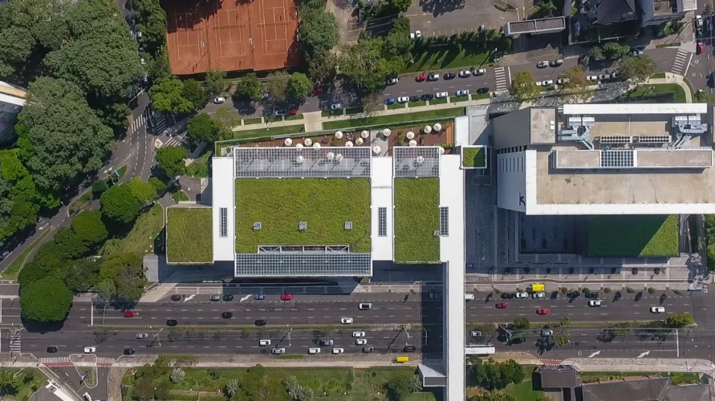 Fotografia aérea vertical de grande escala (vista de drone) capturando um complexo de edifícios corporativos ou institucionais integrados à malha urbana. No centro da imagem, o teto de uma grande edificação branca destaca-se por possuir extensos sistemas de telhado verde, divididos em três grandes blocos retangulares cobertos por uma grama densa e uniforme de cor verde-viva. Estruturas quadriculadas cinzas, possivelmente claraboias ou painéis solares, emolduram as bordas dessas coberturas vegetadas.
O complexo é circundado por avenidas movimentadas com carros em trânsito, calçadas, linhas de arborização urbana densa à esquerda e quadras de tênis de saibro alaranjado no topo. À direita, outra ala do edifício exibe uma área de convivência ao ar livre com ombrelones brancos e pequenos canteiros. A imagem ilustra de forma clara o panorama abordado no artigo sobre a bioarquitetura no Brasil, demonstrando como grandes centros urbanos utilizam tecnologias de infraestrutura verde para atuar como infraestruturas multifuncionais, reduzindo as ilhas de calor, gerenciando as águas pluviais e minimizando os custos operacionais das edificações em áreas com alto índice de pavimentação.