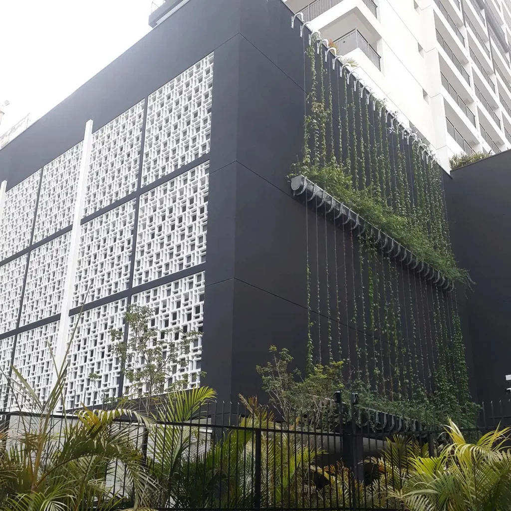 Fachada contemporânea de um edifício de alto padrão pintada em cinza escuro, com painéis de cobogós brancos. À direita, filamentos verticais de um brise vegetal com plantas pendentes descem a partir de uma calha metálica superior, demonstrando a integração da engenharia de fachadas da Ecotelhado para eficiência energética e valorização de ativos ESG.
