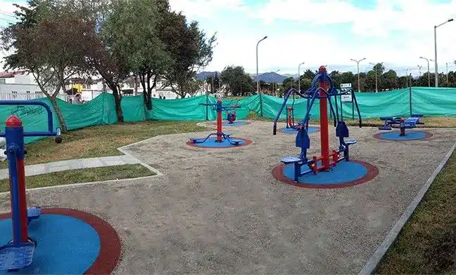Fotografia de uma academia ao ar livre instalada em uma praça ou parque público urbano. O piso de toda a área de circulação é composto pelo sistema de Ecopavimento com preenchimento mineral cinza-claro, delimitado por bordas de concreto que o separam do gramado lateral. Dispostos sobre o pavimento permeável, encontram-se diversos equipamentos de ginástica pintados nas cores azul e vermelha. Cada aparelho está posicionado sobre uma base circular emborrachada azul com borda vermelha.
Ao fundo e nas laterais da área de exercícios, há árvores de médio porte e uma cerca de lona verde que delimita o espaço. Postes de iluminação pública e o topo de montanhas ou árvores distantes são visíveis sob um céu parcialmente nublado. A imagem ilustra de forma prática o uso da tecnologia em espaços públicos, demonstrando como áreas de lazer urbano podem funcionar como superfícies ativas de drenagem para retenção de água da chuva e regulação do microclima contra as ilhas de calor.