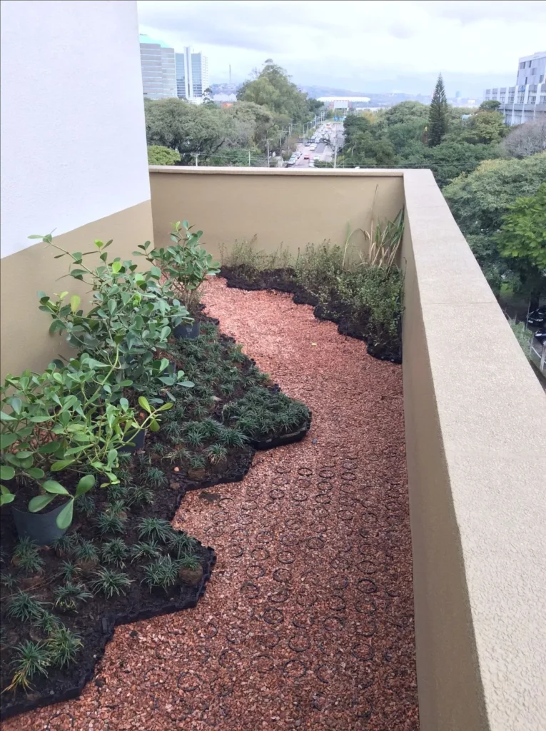 Fotografia em ângulo superior mostrando a sacada ou terraço linear de um edifício residencial ou corporativo, configurado como um teto verde. O piso exibe o sistema de Ecopavimento em duas configurações complementares: um caminho sinuoso preenchido com brita ou pedriscos de tonalidade avermelhada (terracota) e, nas bordas, módulos com revestimento vegetado. À esquerda, mudas de plantas de folhagem verde-escura e pequenos arbustos estão plantados diretamente nos alvéolos estruturais do sistema permeável.
O espaço é delimitado por um parapeito largo de alvenaria pintado em tom bege. Ao fundo da imagem, a vista se abre para uma paisagem urbana arborizada com uma grande avenida movimentada, copas de árvores densas e edifícios comerciais distantes sob um céu nublado. A composição ilustra de forma prática como a infraestrutura verde urbana atua diretamente no combate às ilhas de calor e na promoção da evapotranspiração sobre o solo, equilibrando a temperatura no entorno imediato das edificações.