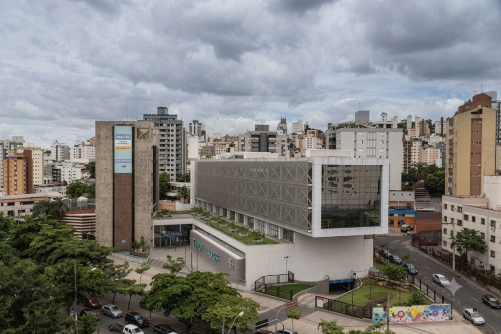 Fotografia aérea parcial de um trecho urbano adensado em Porto Alegre. Em primeiro plano, destaca-se um edifício moderno com telhado verde, representando a aplicação de infraestruturas verdes que o Novo Plano Diretor da cidade busca incentivar. Essa cobertura vegetada serve como uma técnica de prevenção de enchentes por Soluções baseadas na Natureza (SbN), ajudando na retenção e no retardamento do escoamento da água da chuva em áreas urbanas. Ao fundo, vê-se a paisagem consolidada da cidade sob um céu nublado, enfatizando a necessidade de adaptar os espaços existentes.