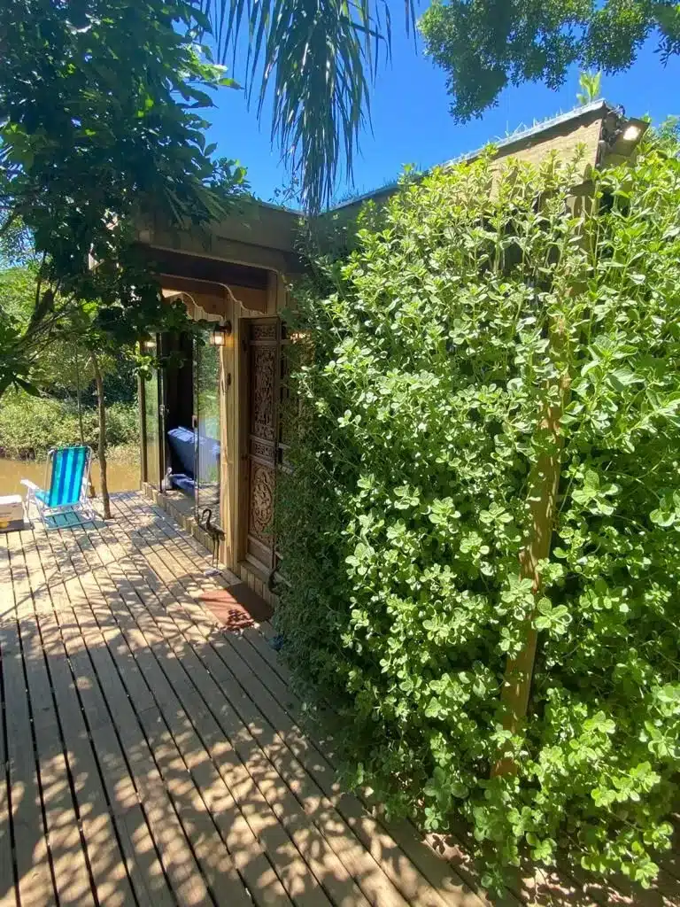 Vista em perspectiva da varanda de uma cabana modular de madeira à beira d'água. O piso é um deck de madeira que projeta sombras das árvores vizinhas. Uma imponente cerca viva de arbustos verdes e folhagens densas cobre a quina da parede direita. Ao fundo, uma porta de madeira esculpida com detalhes ornamentais e uma parede de vidro revelam o interior com um sofá azul. Uma cadeira de praia listrada azul e branca está posicionada no deck voltada para o rio.