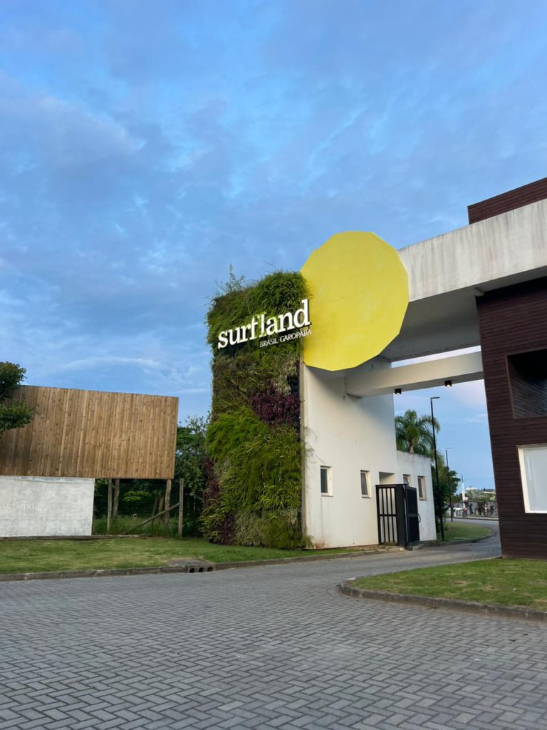 Uma fotografia da entrada principal do Surfland Brasil em Garopaba, Santa Catarina. À esquerda, um grande totem retangular é coberto por um Jardim Vertical Mamute Ecotelhado, exibindo uma vegetação densa e variada com folhagens verdes e roxas, reforçando o caráter biofílico do espaço. Letras luminosas brancas formam o logotipo "surfland" no totem, com o texto "BRASIL GAROPABA" em letras menores abaixo. À direita, um grande círculo amarelo irregular se sobrepõe ao totem verde e à estrutura branca do pórtico de entrada, criando um contraste vibrante. Ao fundo, vê-se um portão preto de acesso e palmeiras, com o complexo arquitetônico do resort e o céu nublado ao anoitecer completando a paisagem integrada à natureza.