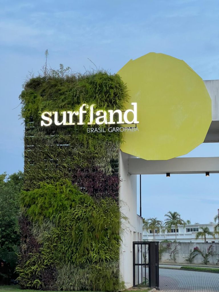 Uma fotografia da entrada principal do Surfland Brasil em Garopaba, Santa Catarina. À esquerda, um grande totem retangular é coberto por um Jardim Vertical Mamute Ecotelhado, exibindo uma vegetação densa e variada com folhagens verdes e roxas, reforçando o caráter biofílico do espaço. Letras luminosas brancas formam o logotipo "surfland" no totem, com o texto "BRASIL GAROPABA" em letras menores abaixo. À direita, um grande círculo amarelo irregular se sobrepõe ao totem verde e à estrutura branca do pórtico de entrada, criando um contraste vibrante. Ao fundo, vê-se um portão preto de acesso e palmeiras, com o complexo arquitetônico do resort e o céu nublado ao anoitecer completando a paisagem integrada à natureza.