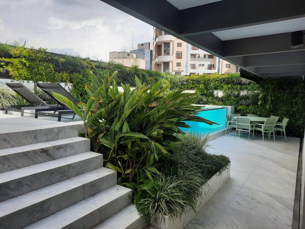 Área de lazer ao ar livre em uma cobertura moderna, com escada de mármore claro no primeiro plano, canteiro com plantas tropicais densas, piscina com parede de vidro transparente e espreguiçadeiras. Ao fundo, uma mesa de jantar externa sob pérgula e prédios urbanos sob céu nublado.