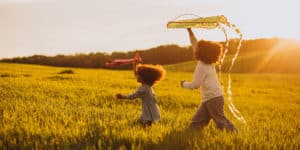 Imagem de duas irmãs correndo uma atrás da outra, brincando de pipa, em um campo, para simbolizar a saúde mental infantil