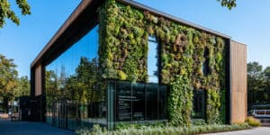 Edifício moderno com fachada de vidro e parede viva coberta por plantas, refletindo árvores ao redor em um dia ensolarado, simbolizando arquitetura sustentável e integração com a natureza.