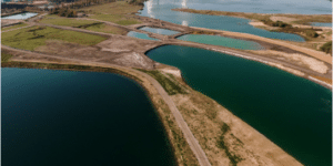 Vista aérea de um sistema de lagoas artificiais com diferentes tonalidades de água, rodeado por áreas de terra escavada e vegetação rala, possivelmente relacionado a um processo de tratamento hídrico ou contenção industrial, com instalações industriais visíveis ao fundo.