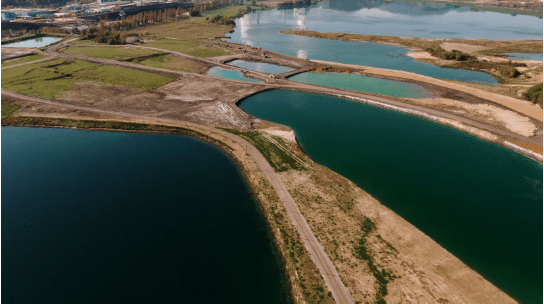Vista aérea de um sistema de lagoas artificiais com diferentes tonalidades de água, rodeado por áreas de terra escavada e vegetação rala, possivelmente relacionado a um processo de tratamento hídrico ou contenção industrial, com instalações industriais visíveis ao fundo.