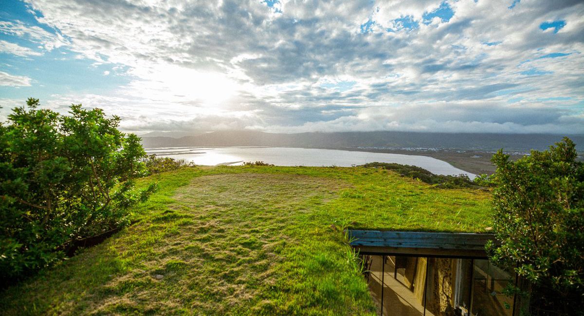 Vista deslumbrante de uma paisagem natural ao pôr do sol, com vegetação exuberante e telhado verde integrando-se ao ambiente; na parte inferior direita, estrutura arquitetônica moderna com uso de vidro e elementos sustentáveis, ideal para projetos que valorizam soluções ecológicas como telhados verdes e jardins verticais artificiais.