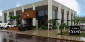 Fachada moderna da loja Elevato Casa em Xangri-Lá, apresentando uma arquitetura contemporânea com amplas vidraças, colunas brancas estruturais e detalhes em madeira. O ambiente externo exibe um paisagismo integrado com árvores ornamentais e arbustos, exemplificando o conceito de design biofílico e sustentável mencionado no projeto, que utiliza jardins verticais e elementos naturais para promover conforto térmico e purificação do ar. O chão está molhado, refletindo a estrutura em um dia de chuva.