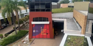 Vista de cima do pátio central do Paseo Zona Sul Shopping em Porto Alegre, destacando uma estrutura arquitetônica central de cor terracota com um grande banner roxo onde se lê "É muito bom ser daqui". A imagem exemplifica o conceito aberto e biofílico do empreendimento, mostrando, em diferentes níveis, Telhados Verdes da Ecotelhado, um espelho d'água com pedras e palmeiras altas, reforçando a integração entre soluções sustentáveis e arquitetura contemporânea para convivência ao ar livre.
