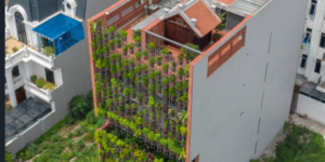 Vista aérea oblíqua de um edifício residencial urbano estreito e alto com uma fachada funcional de agritetura. A face frontal da construção é inteiramente coberta por um jardim vertical composto por dezenas de pequenos vasos suspensos com plantas e hortaliças verdes, criando uma parede viva texturizada. No topo do prédio, há um terraço com telhas cerâmicas avermelhadas e uma pequena estrutura de madeira no estilo tradicional asiático.