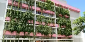 A imagem mostra um prédio universitário nas cores branco e rosa com brises verticais se estendendo na fachada, criando uma atmosfera que harmoniza o urbano com o natural.